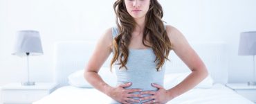 Young woman sat on a bed clutching her stomach to represent tummy ache